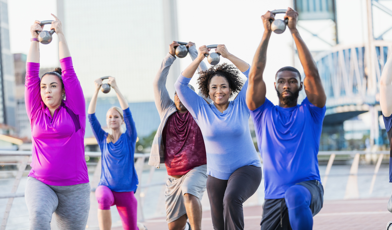 A mid adult African-American man in his 30s, wearing a blue shirt, leading a multi-ethnic exercise class of mixed ages. The oldest in the group is a senior woman in her 60s, in the middle next to the instructor, smiling at the camera. They are lifting kettle bells above their heads, doing lunges. They are outdoors on Jacksonville's Riverfront 